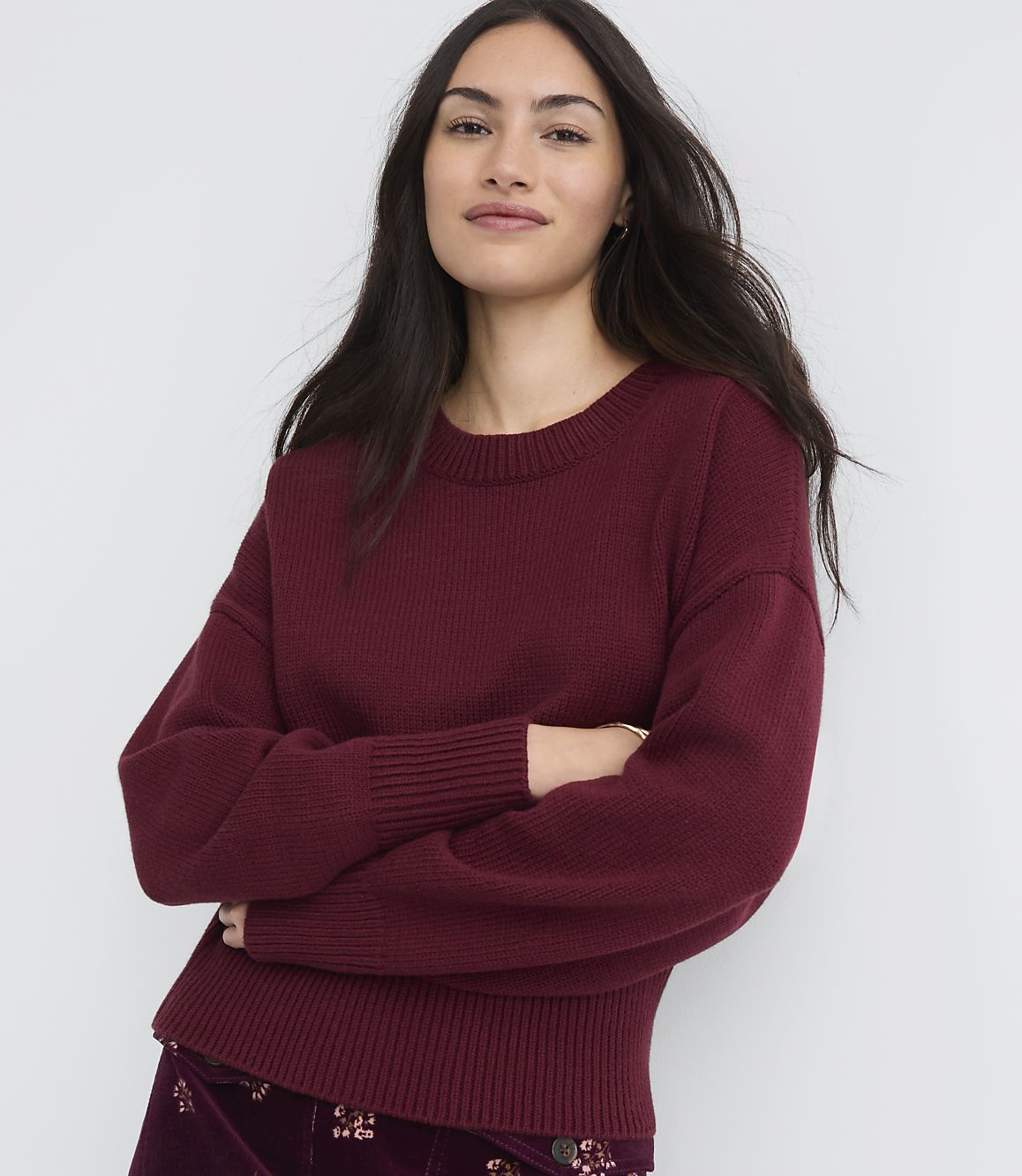 A woman in a maroon sweater and patterned skirt stands with her arms crossed against a plain white background.