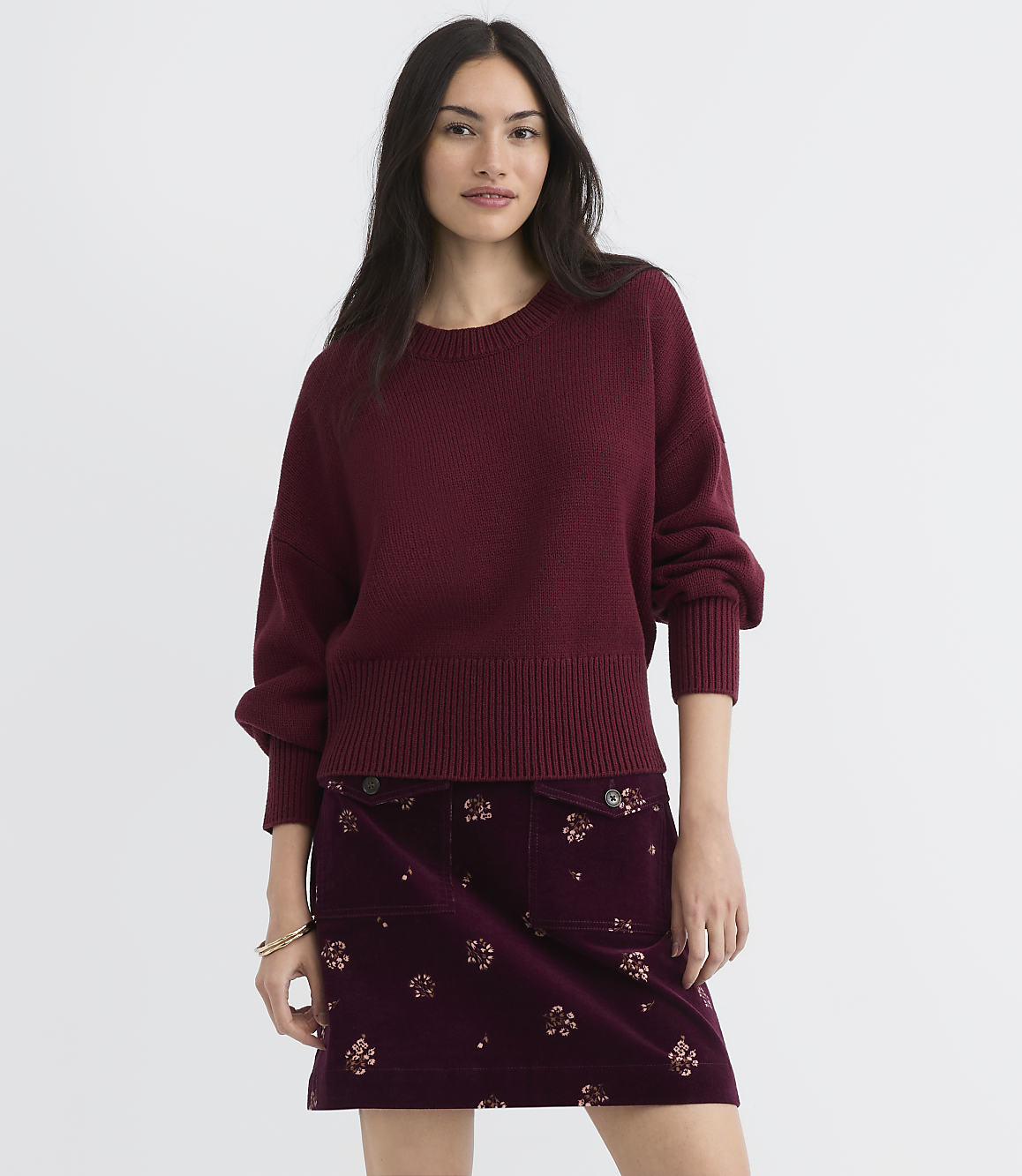 A woman in a maroon sweater and floral skirt stands against a plain white background.