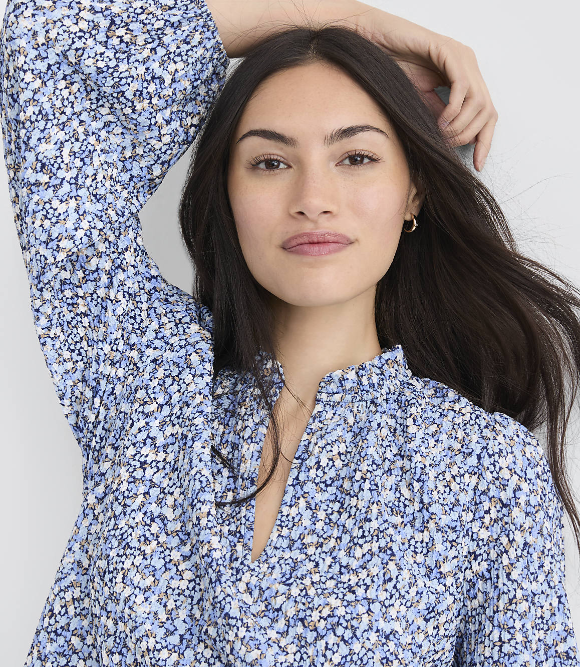 A woman with long dark hair, wearing a blue floral print dress, poses with her arm raised against a plain white background.