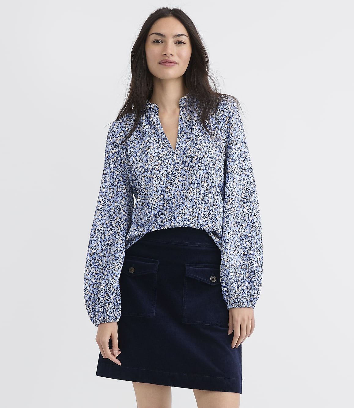 A woman in a floral blouse and dark skirt stands against a plain white background.