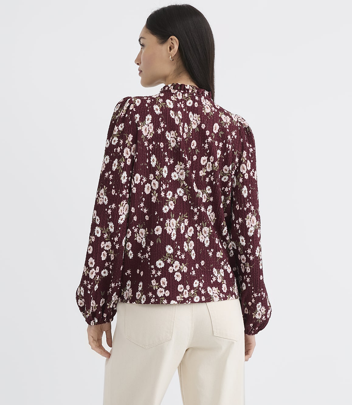 A woman in a floral blouse and light-colored pants stands against a plain white background.