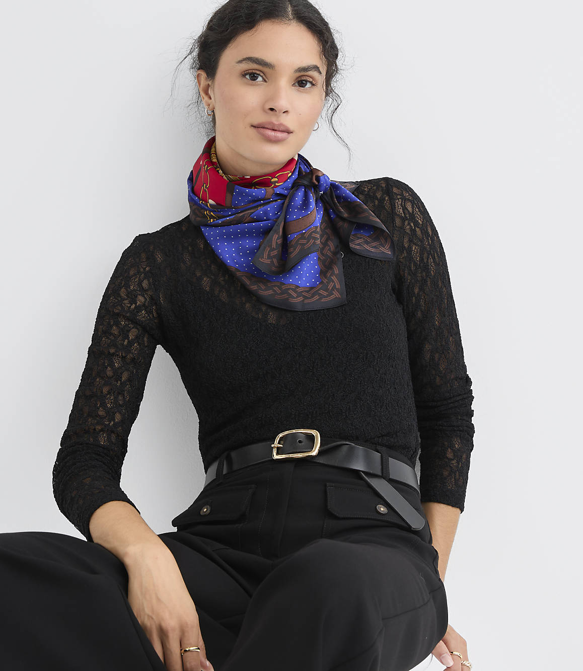 A woman in a black lace top and black pants with a patterned scarf tied around her neck sits against a plain white background.