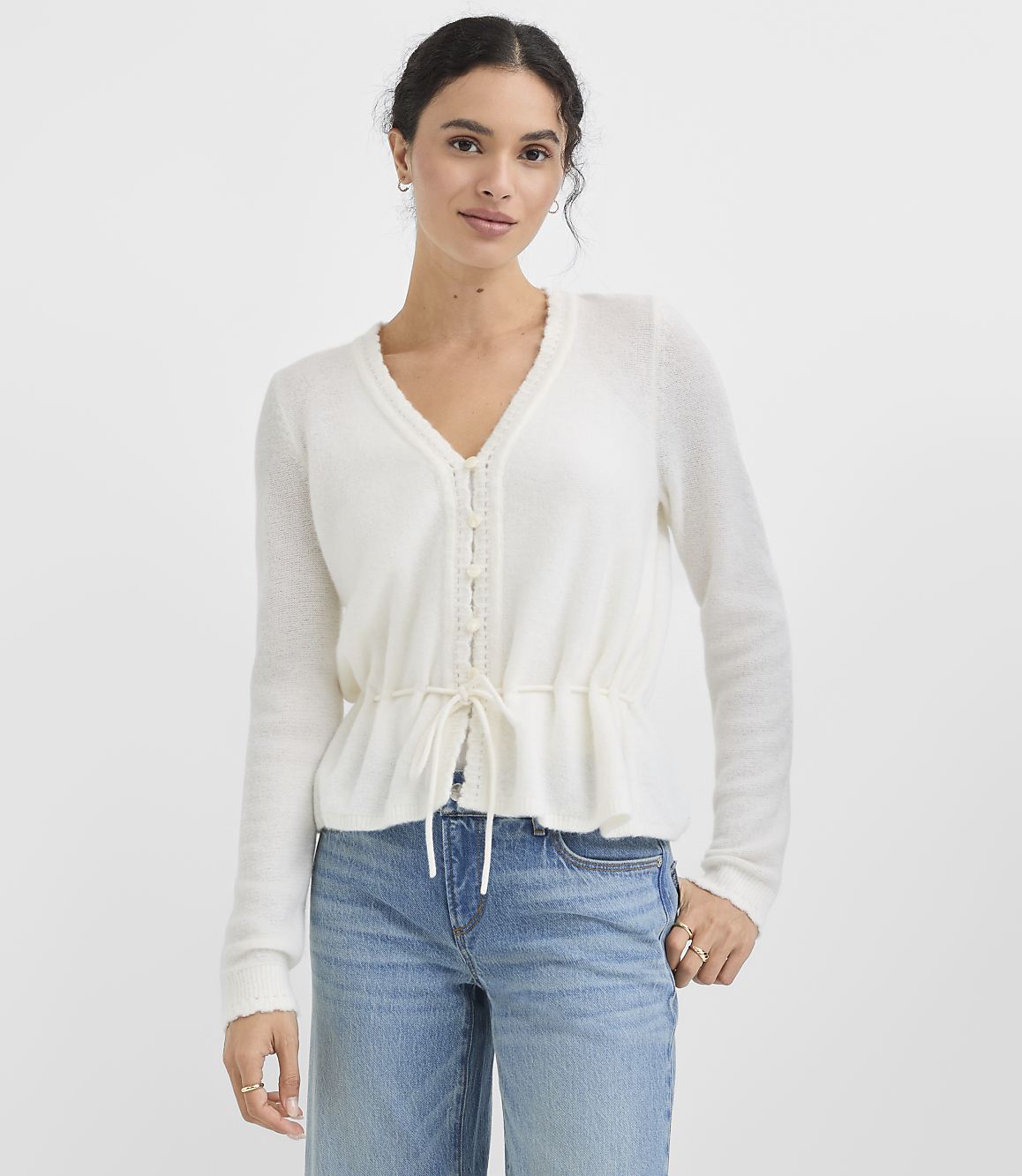 A woman in a white cardigan and blue jeans stands against a plain white background.