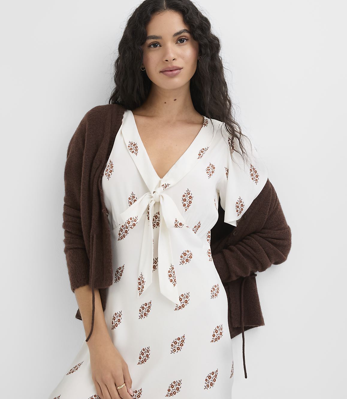 A woman in a white floral dress and brown cardigan stands against a plain white background.