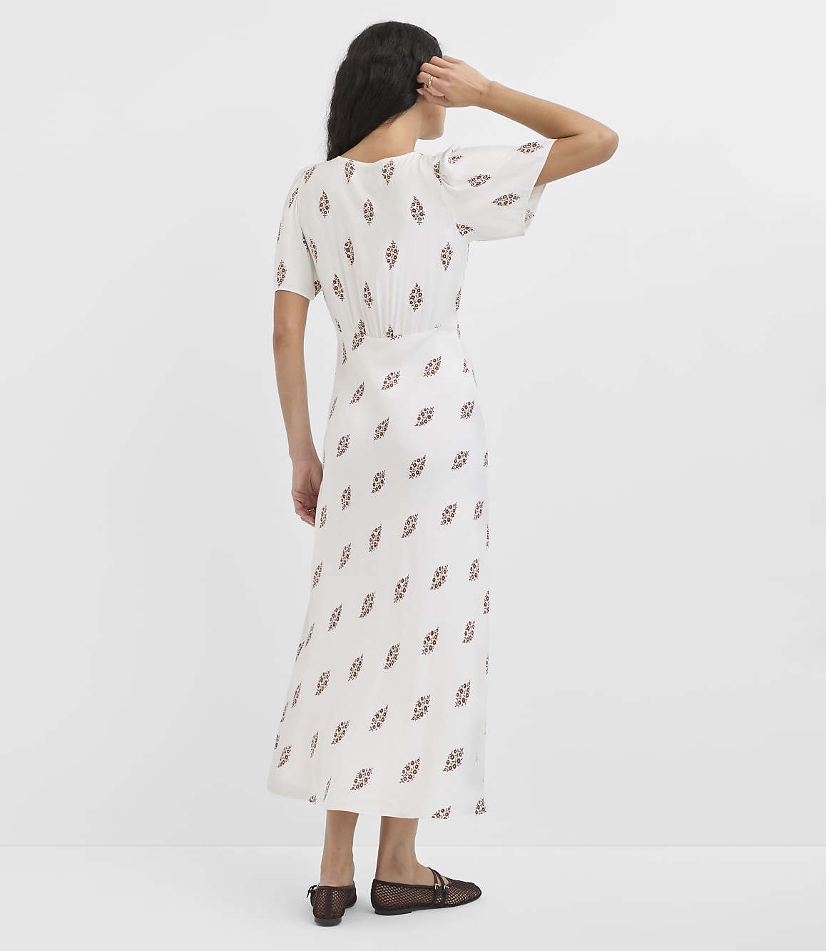 A woman in a white, patterned dress stands against a plain white background.