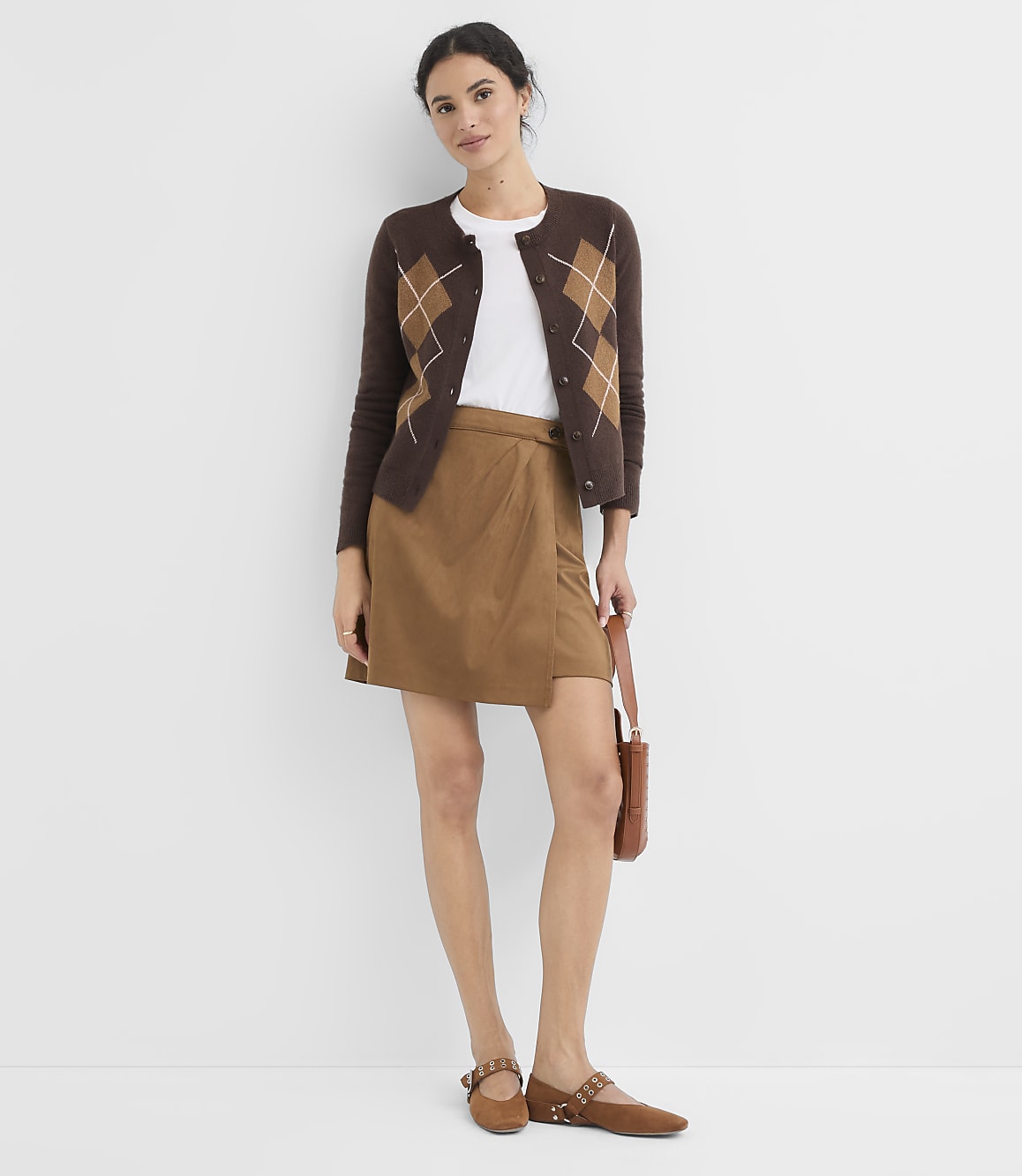 A woman in a brown argyle cardigan and tan skirt stands against a plain white background, holding a brown purse.