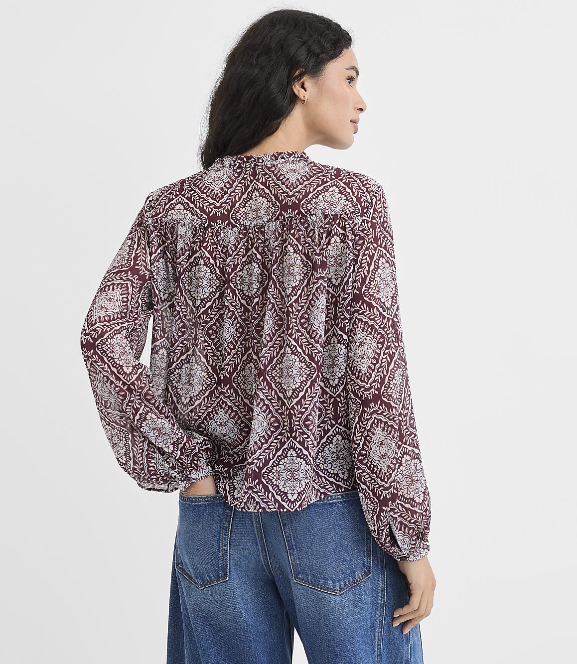 A woman in a patterned maroon blouse and blue jeans stands with her back to the viewer against a plain white background.