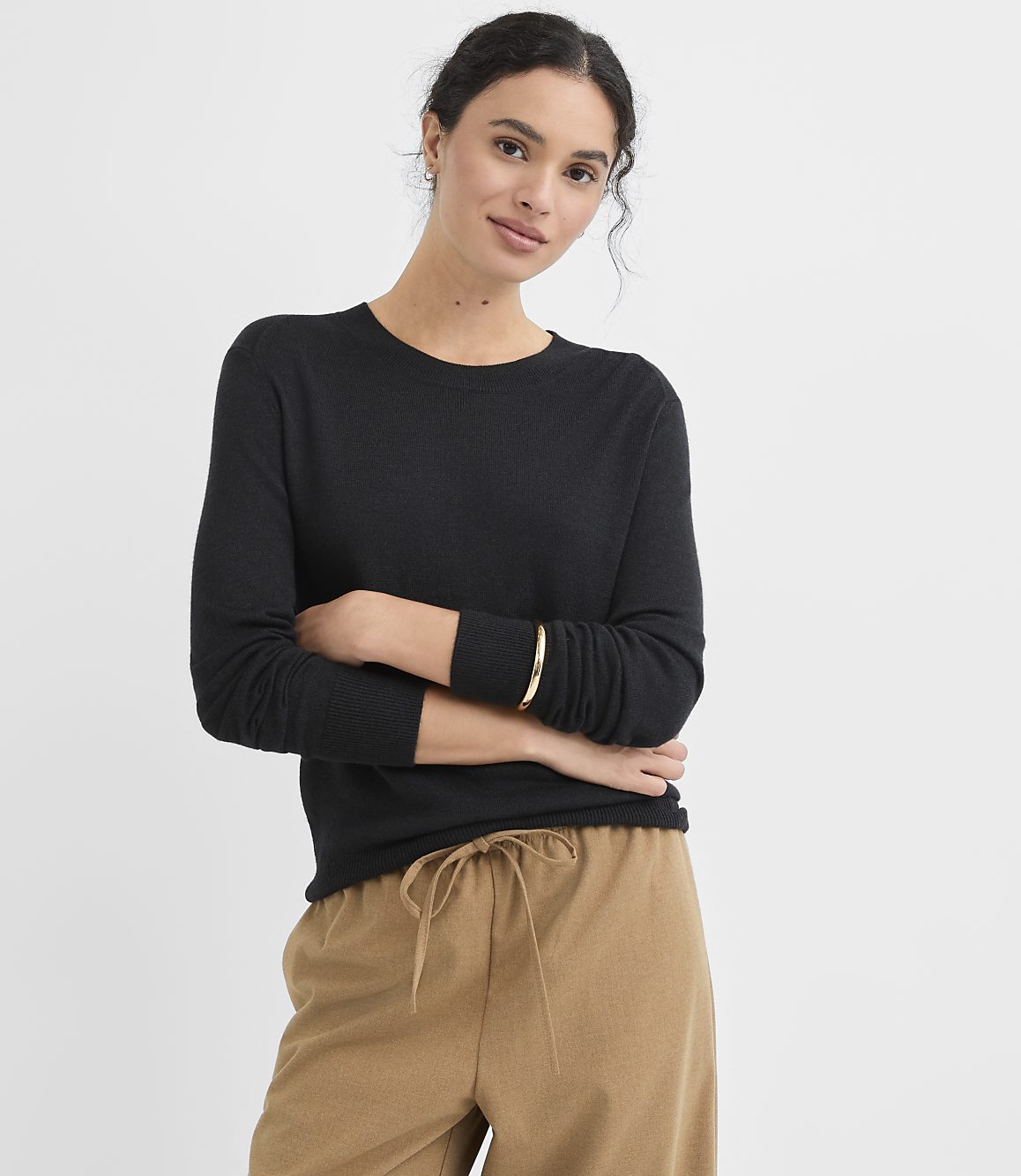 A woman in a black sweater and tan pants stands against a plain white background.