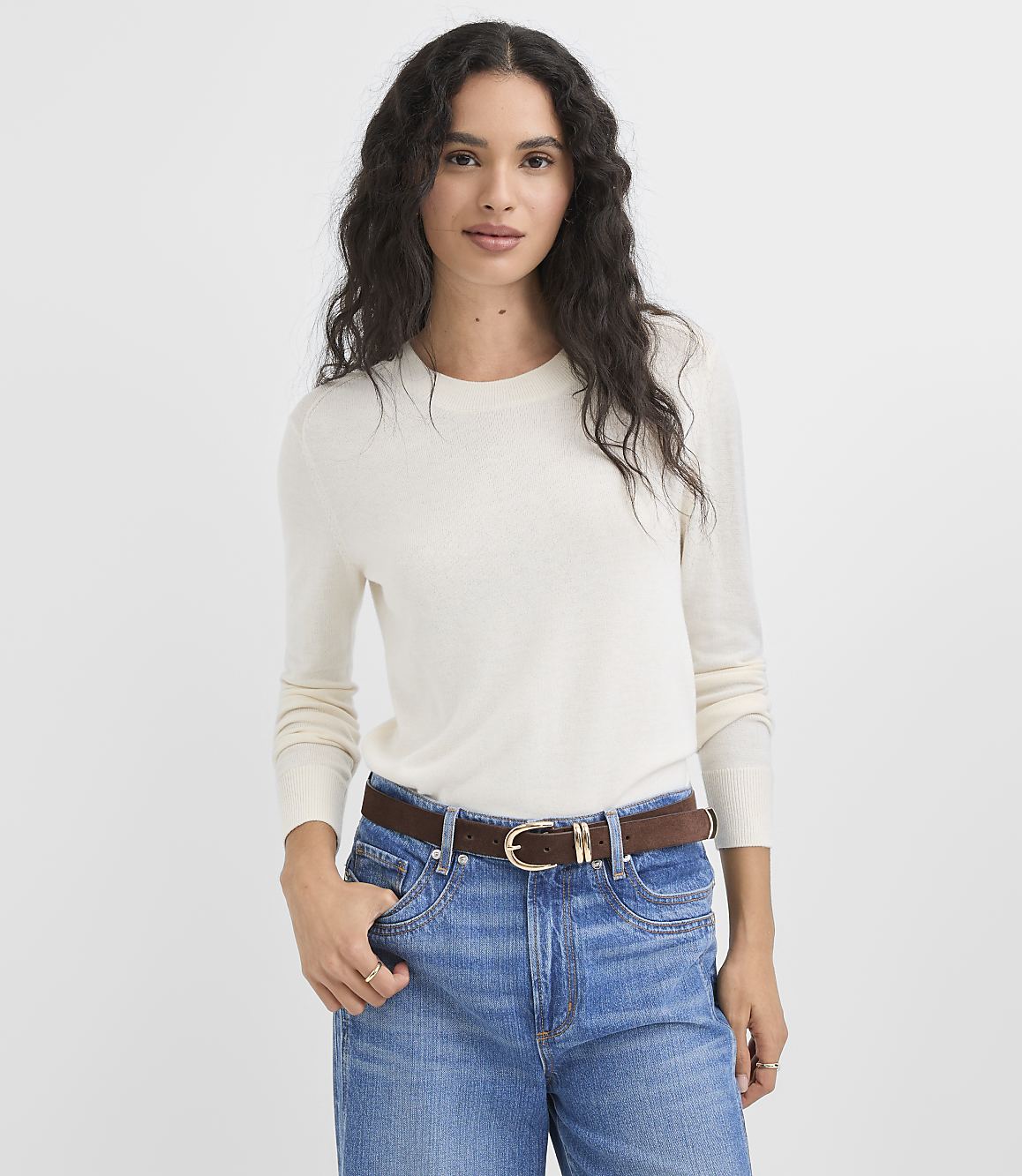 A woman in a cream sweater and blue jeans stands against a plain white background.