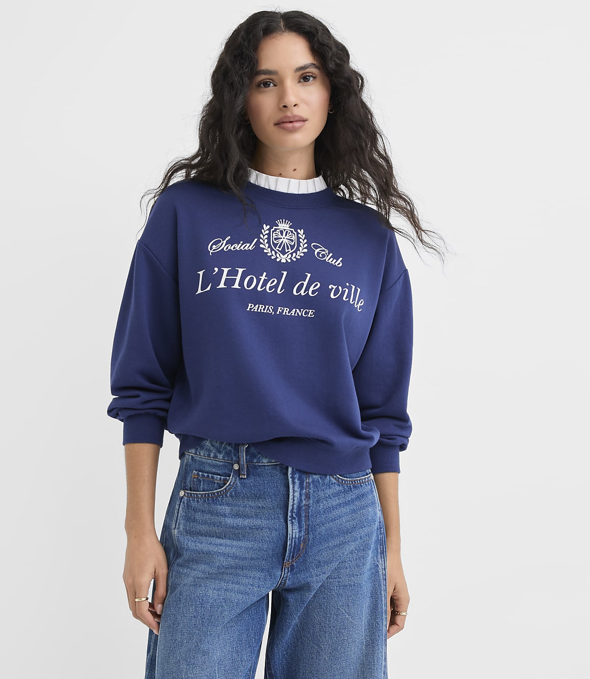 A woman in a blue sweatshirt and jeans stands against a plain white background.