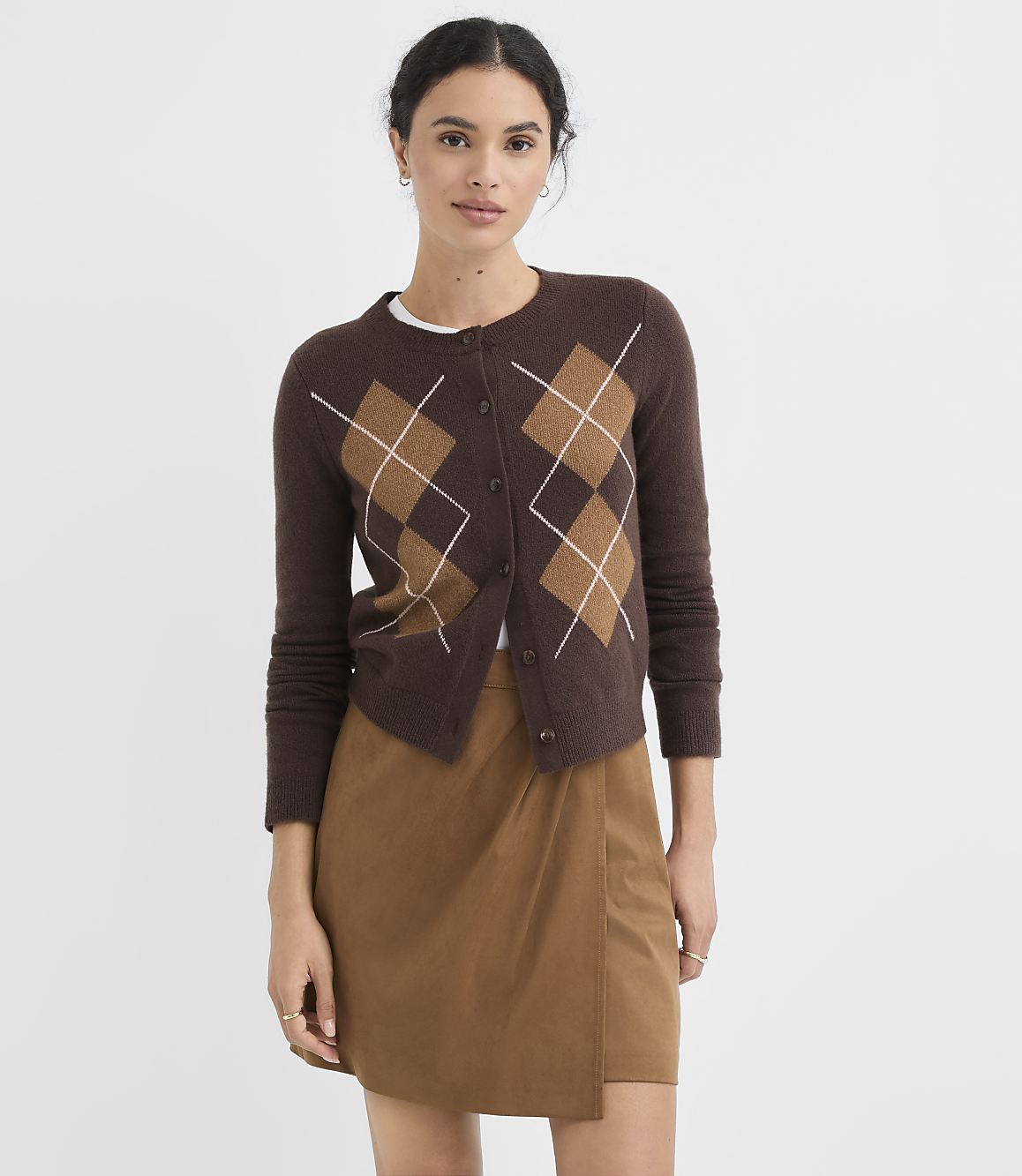 A woman in a brown argyle cardigan and a tan wrap skirt stands against a plain white background.
