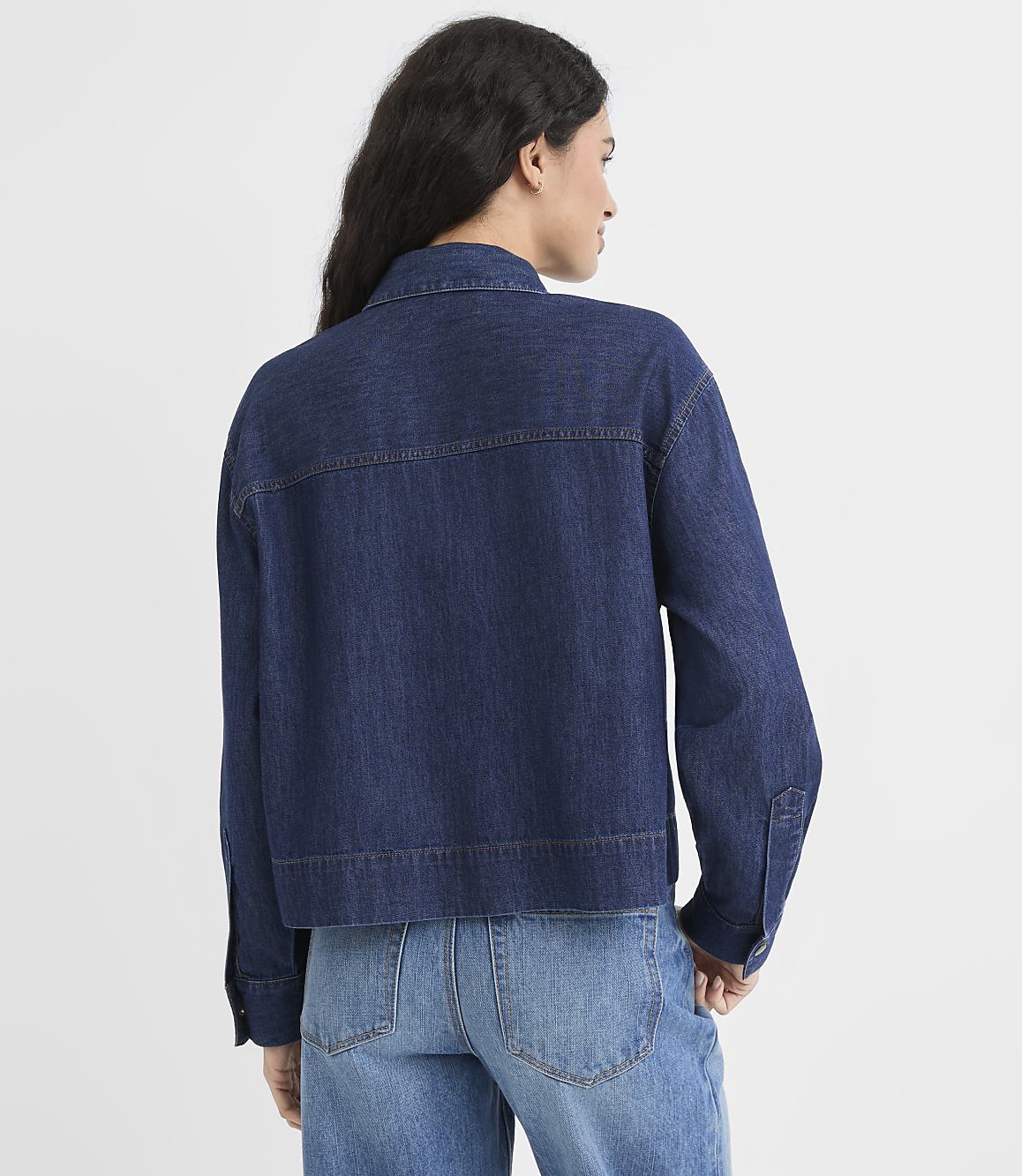 A woman in a denim jacket and jeans stands against a plain white background.