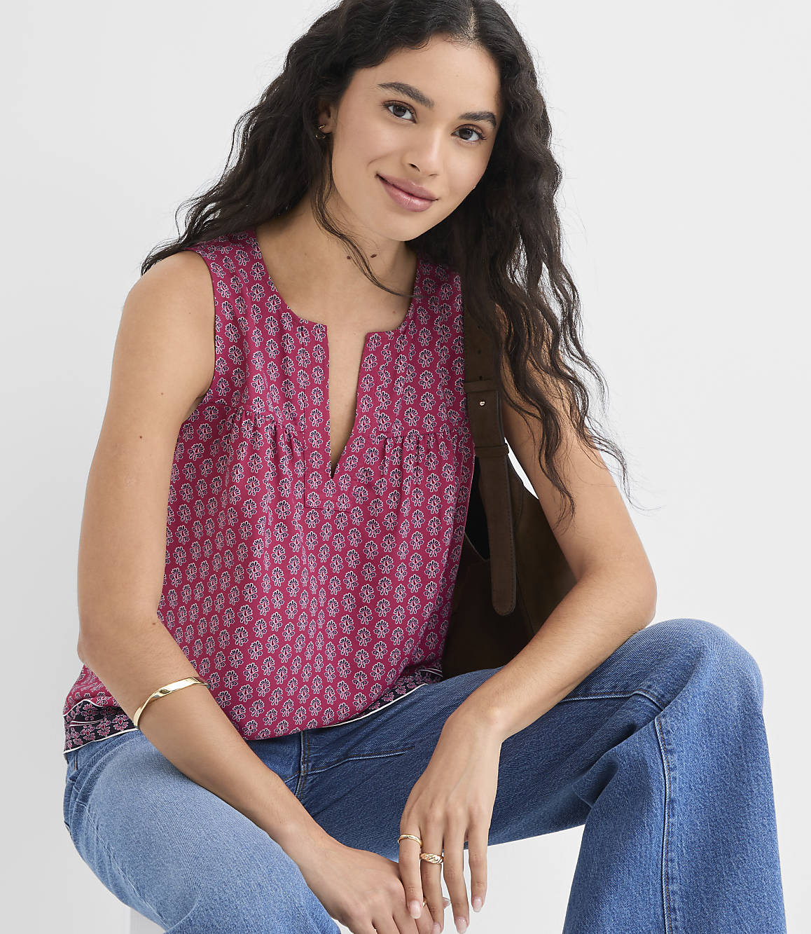 A woman with long dark hair wearing a pink patterned sleeveless top and blue jeans sits with a brown bag over her shoulder against a plain background.