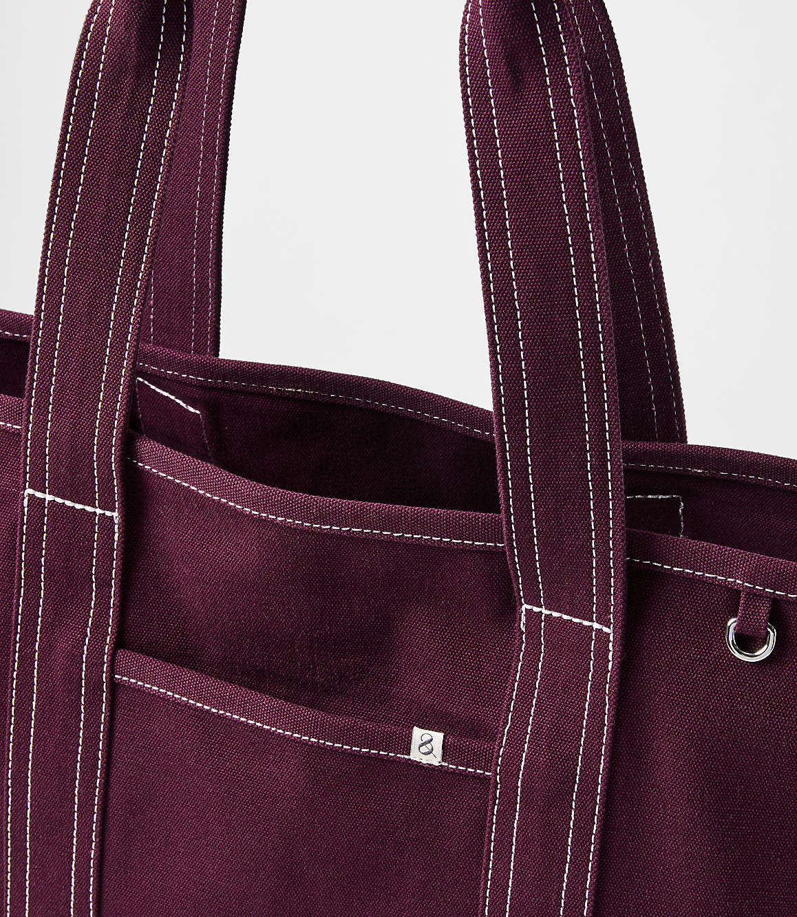 A close-up of a burgundy canvas tote bag with white stitching and a small logo is presented against a plain white background.