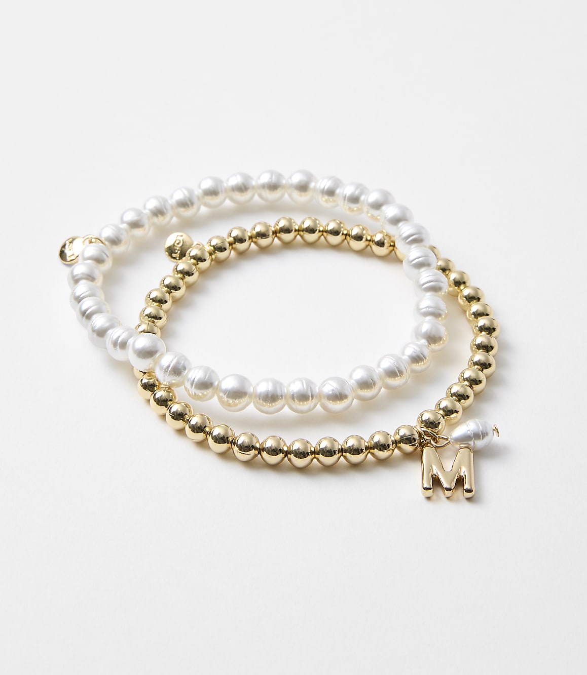 Two bracelets, one with pearls and the other with gold beads and an "M" charm, rest on a plain white background.