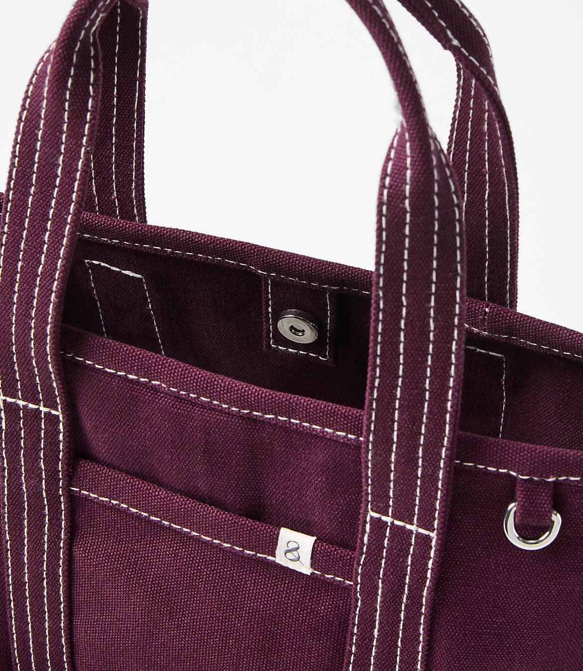 A close-up of a burgundy canvas tote bag with white stitching and a silver snap closure is set against a plain white background.