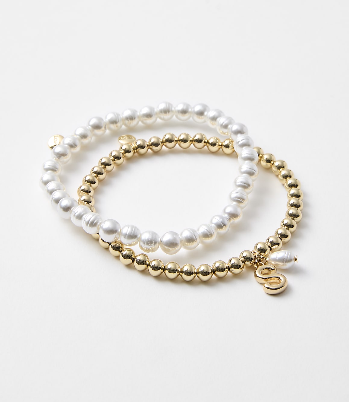 Two bracelets, one with pearls and the other with gold beads and an "S" charm, rest on a plain white background.