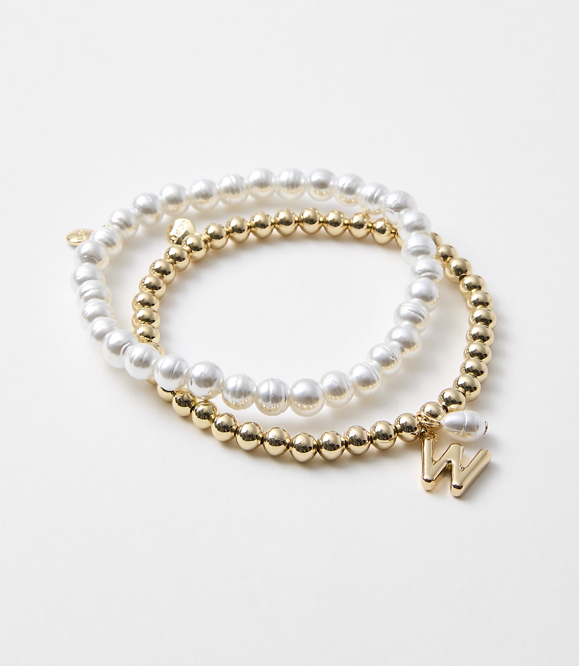 Two bracelets, one with white pearls and the other with gold beads and a gold "W" charm, rest on a plain white background.