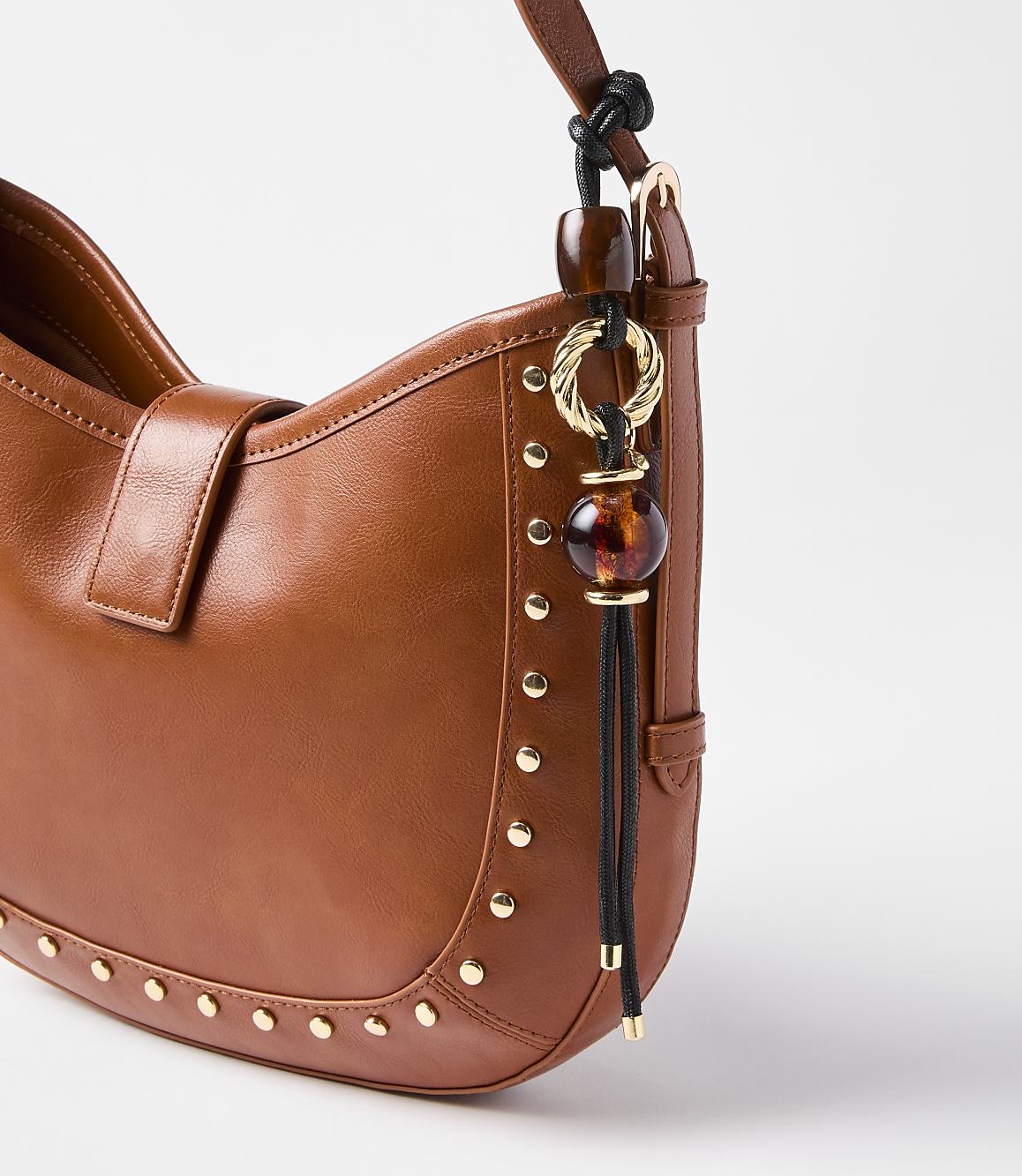 A close-up of a brown leather handbag with gold stud detailing and a decorative charm hangs against a plain white background.