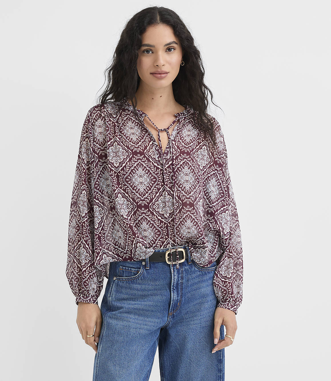A woman in a patterned burgundy blouse and blue jeans stands against a plain white background.