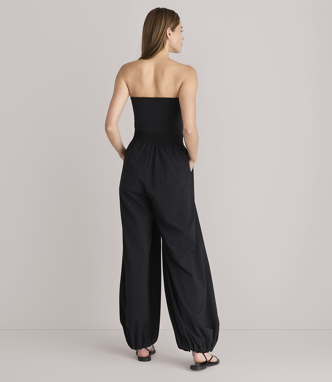 A woman in a black strapless jumpsuit stands with her hands in her pockets against a plain white background.