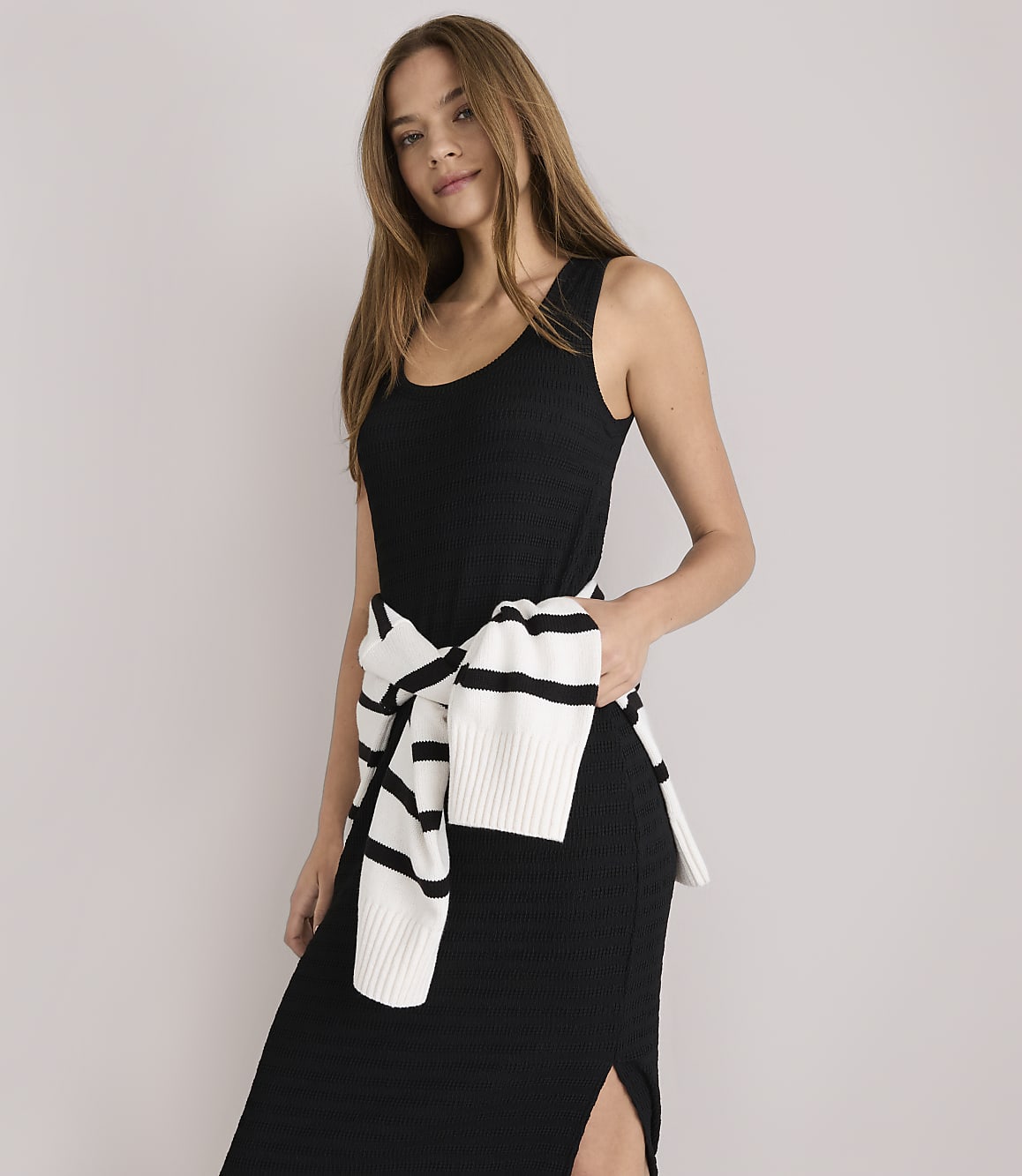 A woman in a black dress with a striped sweater tied around her waist stands against a plain white background.
