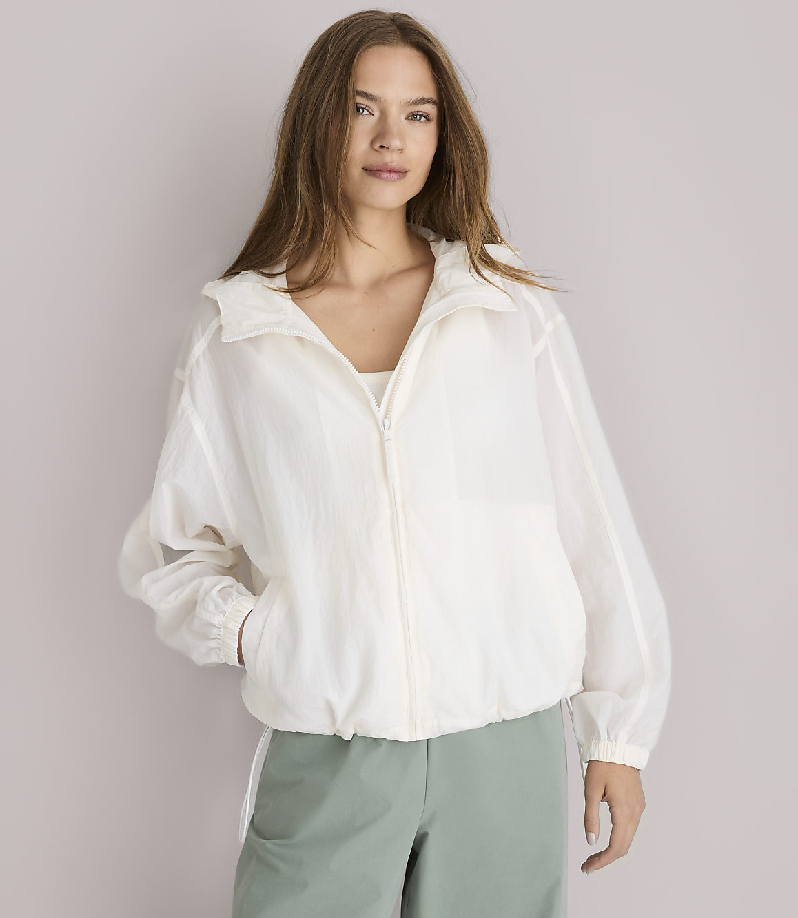 A young woman in a white jacket and green pants stands against a plain white background.