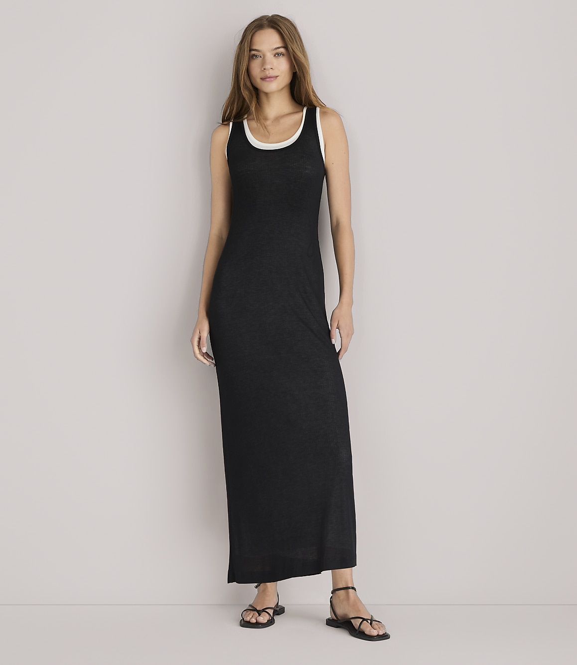 A woman in a black maxi dress and denim jacket leans against a white wall.