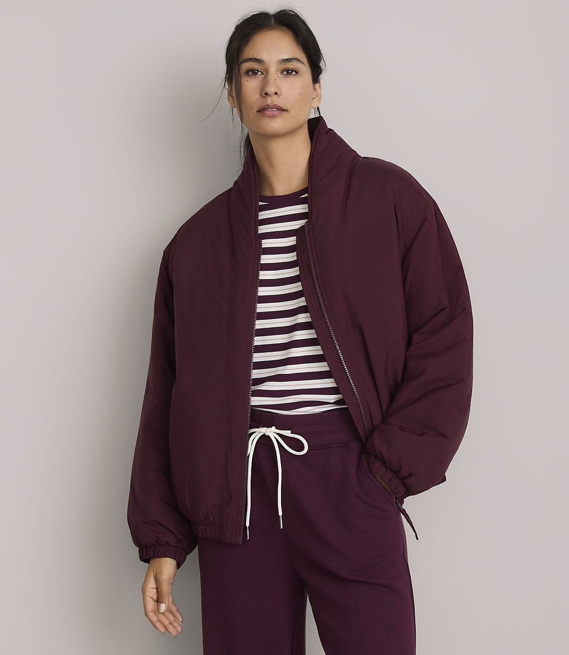 A woman in a burgundy puffer jacket and striped shirt stands against a plain gray background.