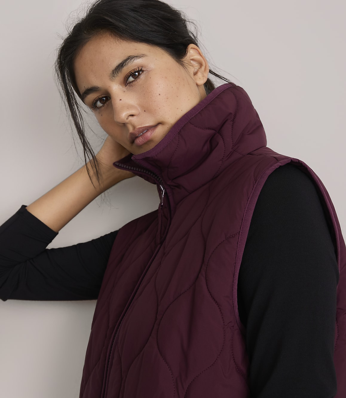 A woman in a burgundy quilted vest and black top poses against a plain, light background.