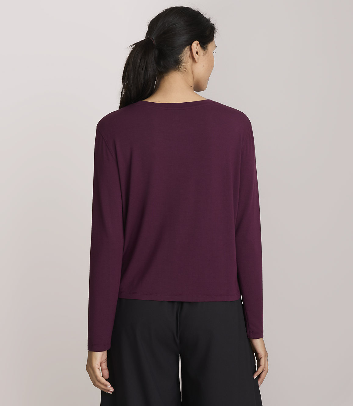 A woman in a maroon long-sleeved shirt and black pants stands against a plain, light purple background.