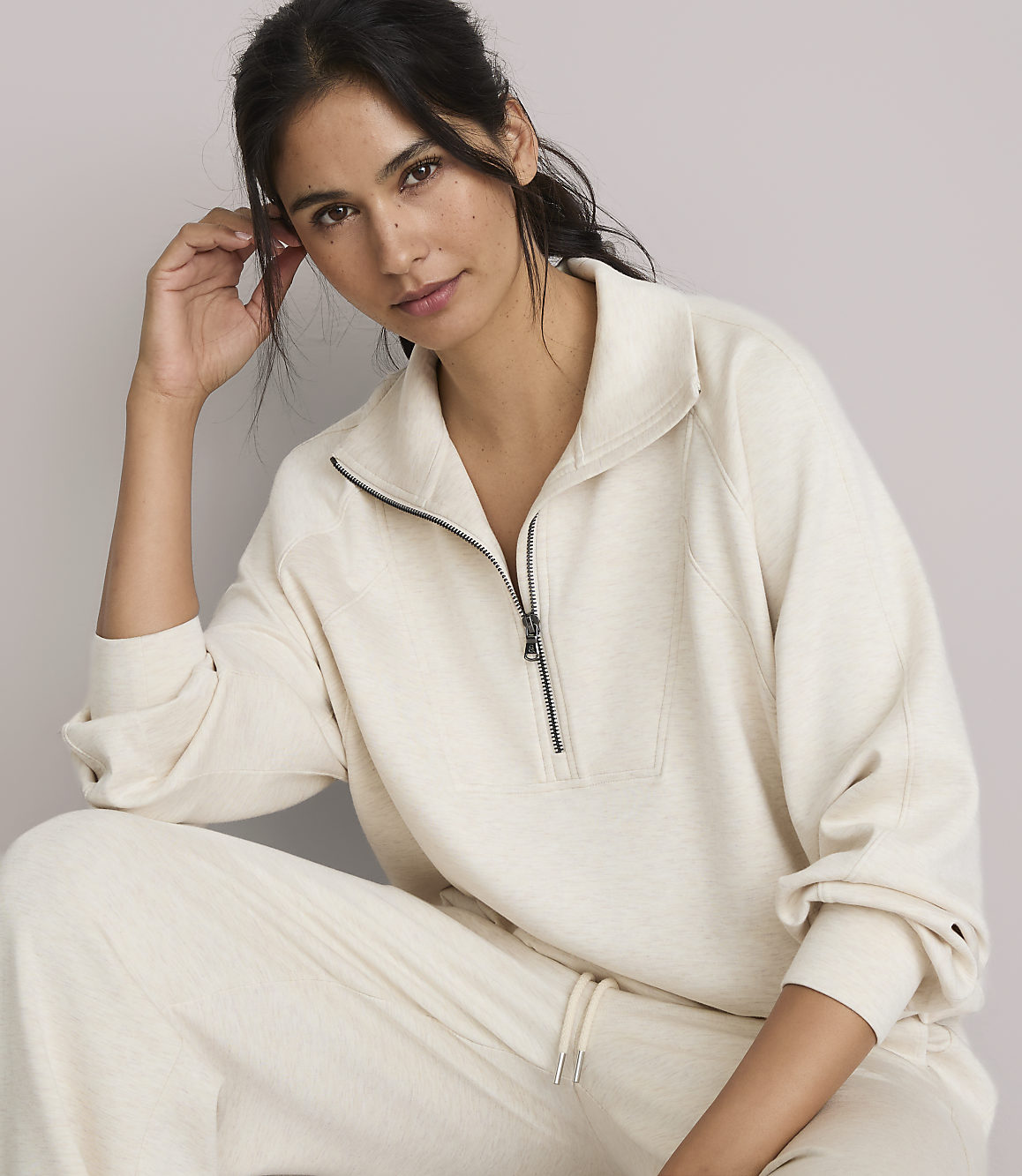 A woman in a cream-colored quarter-zip sweatshirt and matching pants poses against a plain, light gray background.