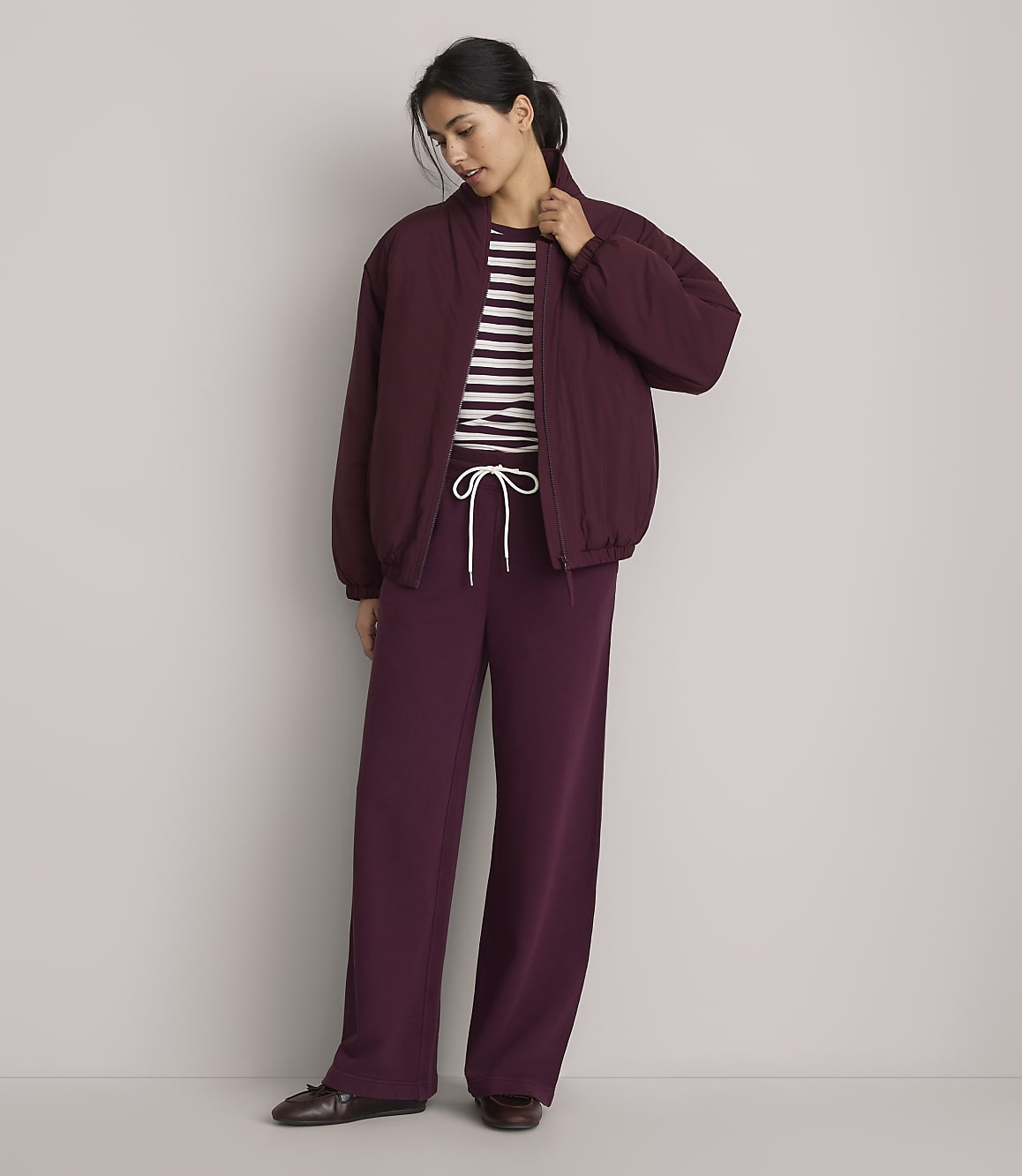 A woman in a burgundy bomber jacket and matching wide-leg pants stands against a plain gray background.