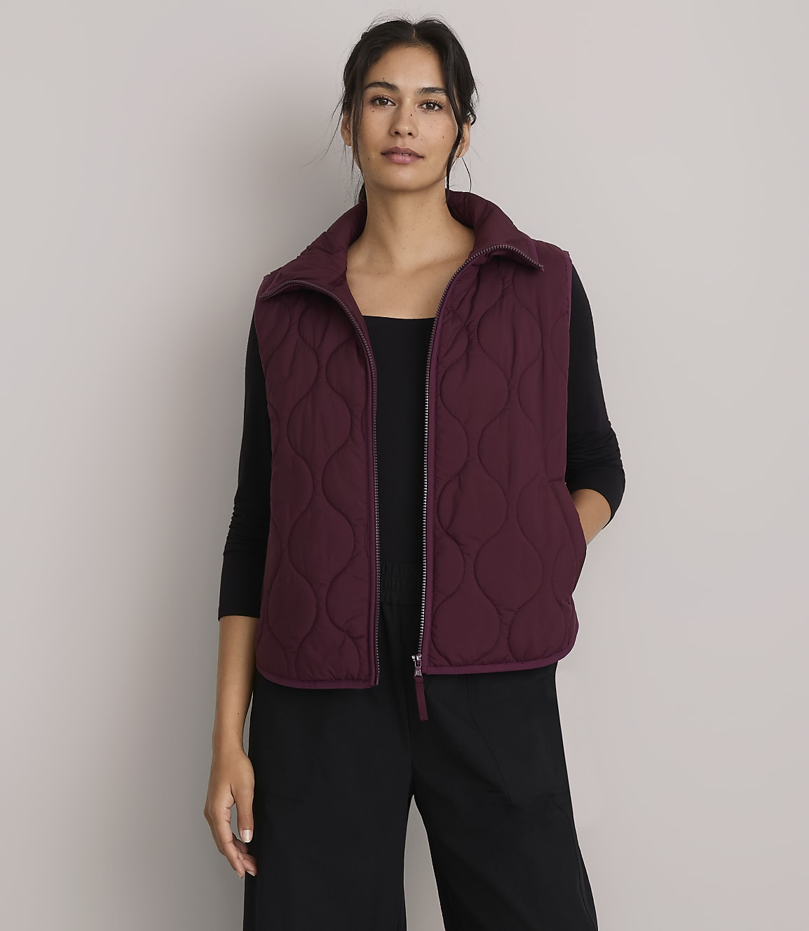 A woman in a quilted vest and hoodie stands against a plain purple background, holding a striped sweater.
