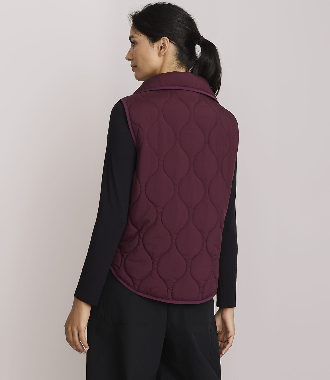 A woman in a quilted burgundy vest and black clothing stands against a plain, light-colored background.