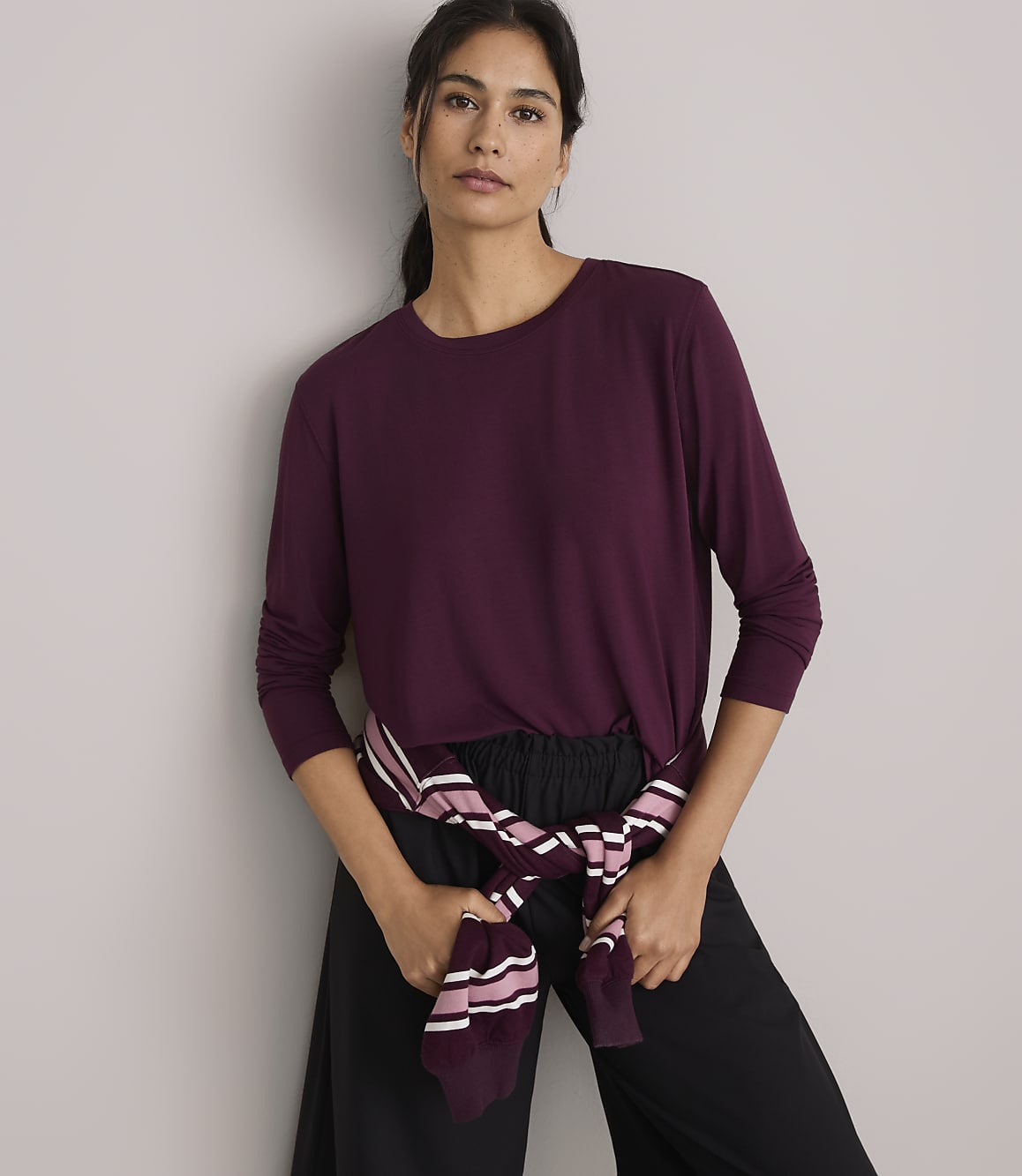 A woman in a burgundy long-sleeved shirt and black pants stands against a plain gray background, with a striped sweater tied around her waist.