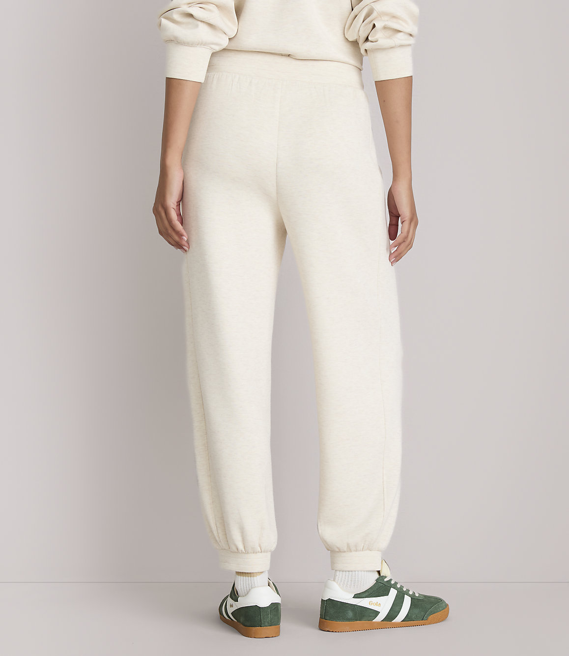 A person wearing cream-colored sweatpants and green and white sneakers stands against a plain, light-colored background.