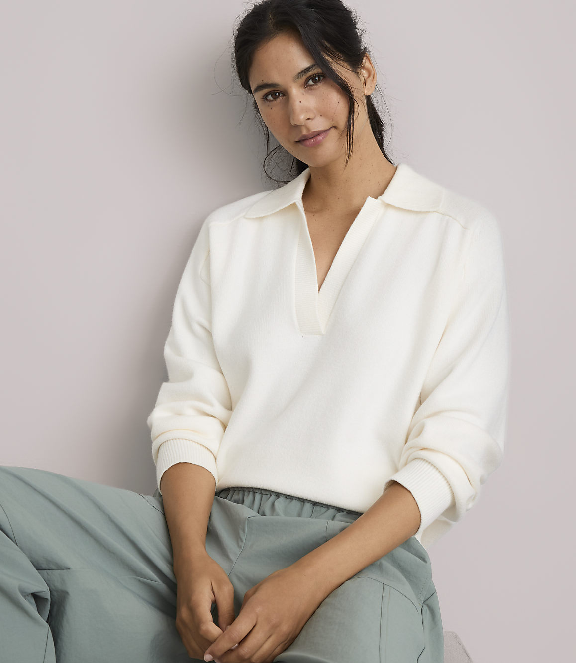 A woman in a cream-colored collared sweater and light green pants sits against a plain, light-colored background.
