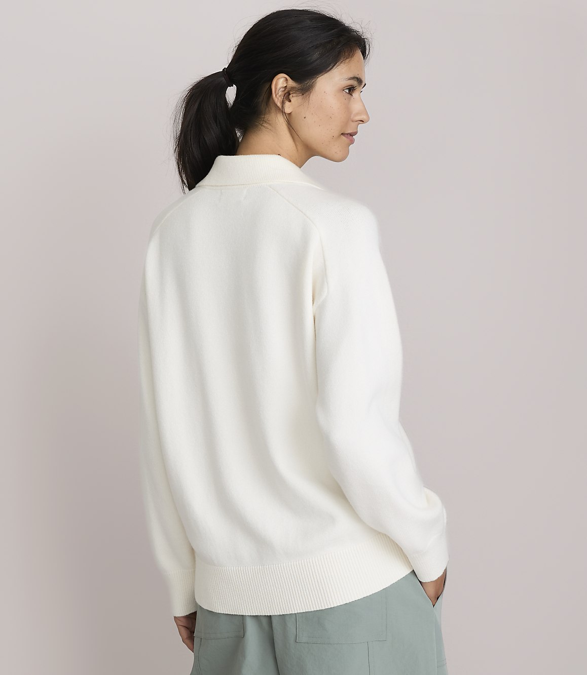 A woman in a cream sweater and light green pants stands against a plain, light pink background.