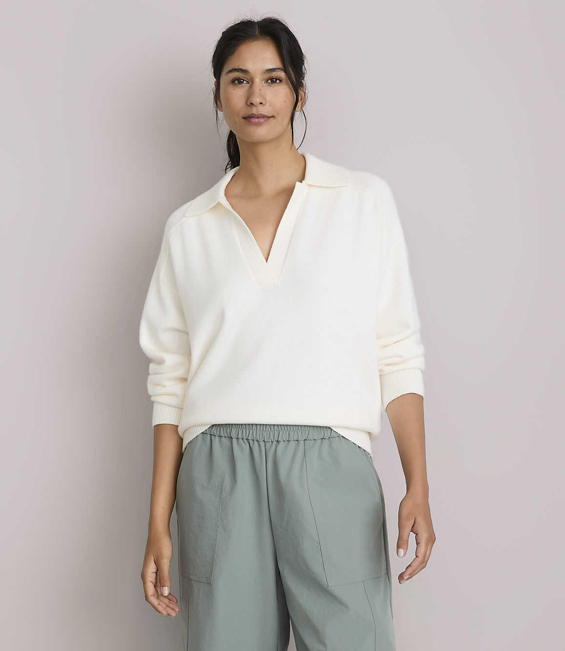 A woman in a cream-colored collared sweater and green pants stands against a plain, light gray background.