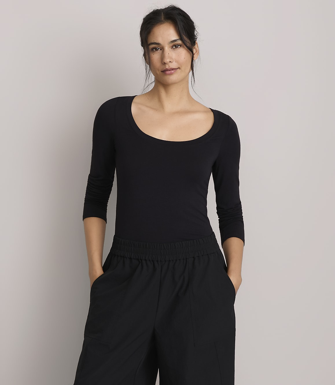 A woman in a black scoop-neck shirt and black pants stands against a plain gray background.