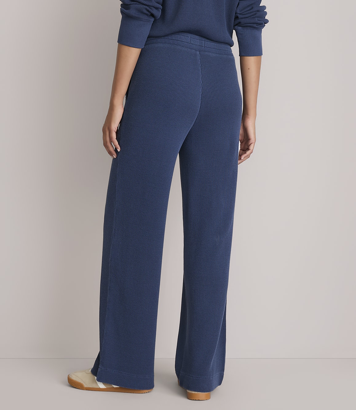 A person wearing blue ribbed lounge pants and cream-colored shoes stands against a plain gray background.