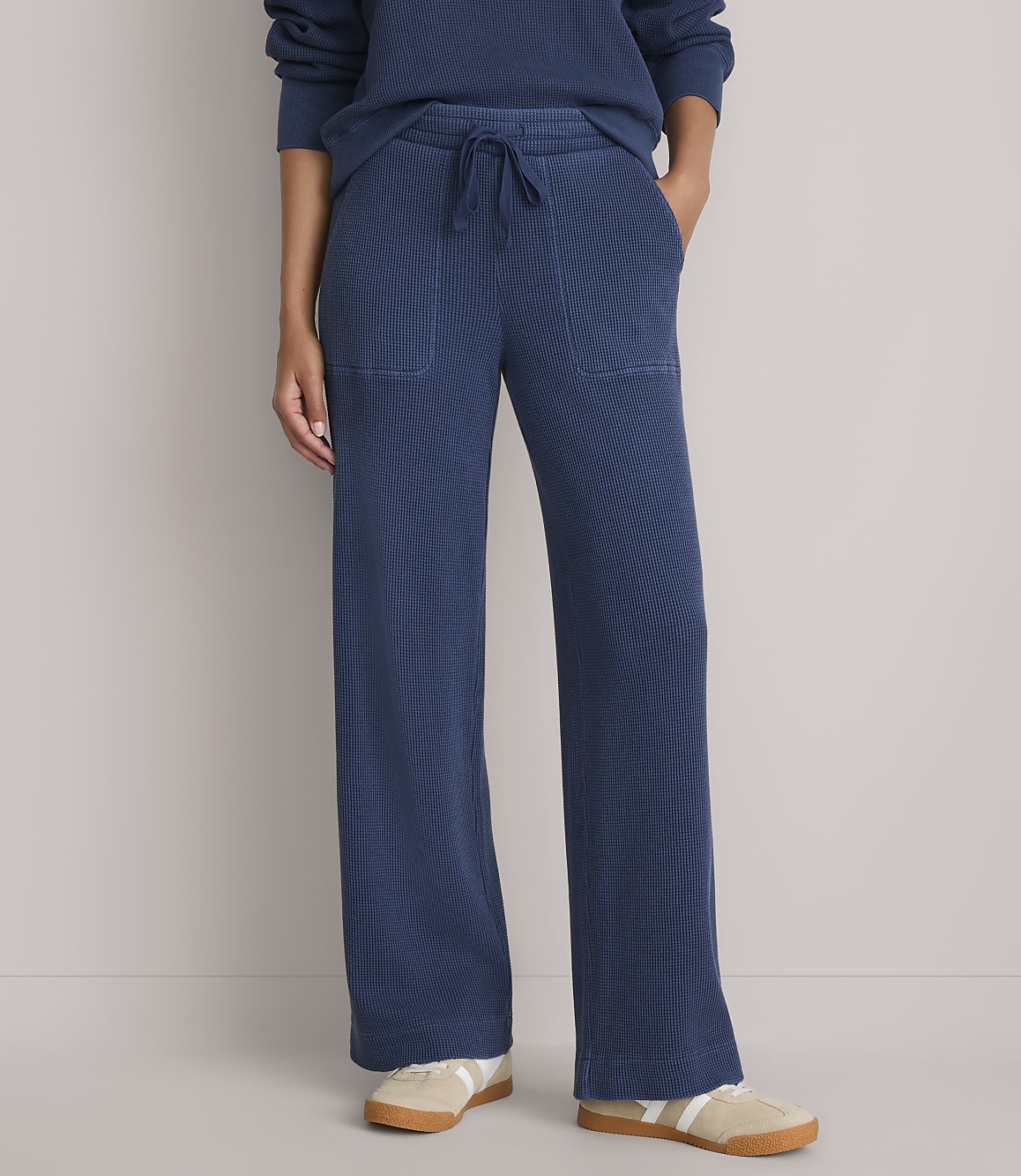 A person stands wearing blue ribbed lounge pants with pockets and a matching top, against a plain grey background.