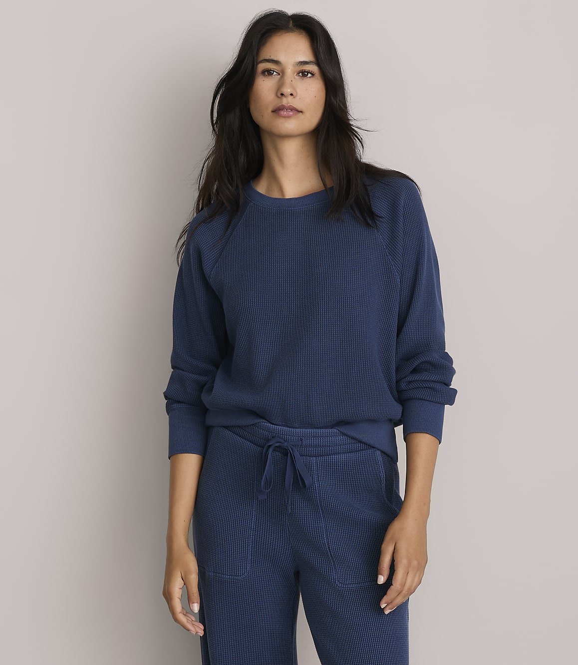 A woman in a blue sweatshirt and matching pants stands against a plain gray background.