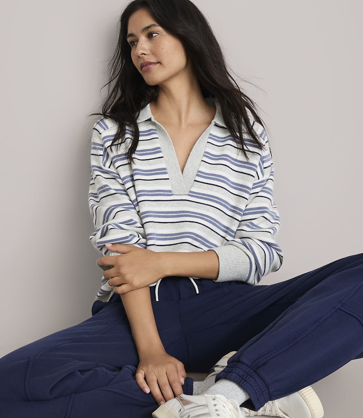 A woman in a striped collared shirt and navy sweatpants sits against a plain, light-colored background.