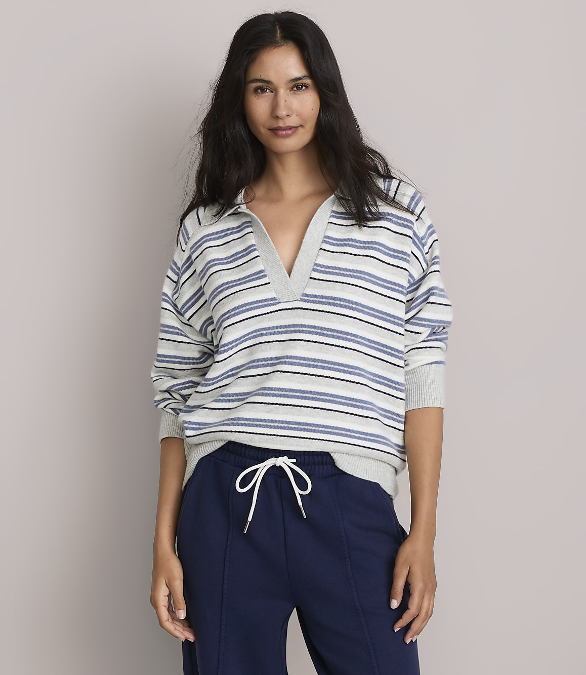 A woman in a striped collared shirt and navy sweatpants stands against a plain gray background.