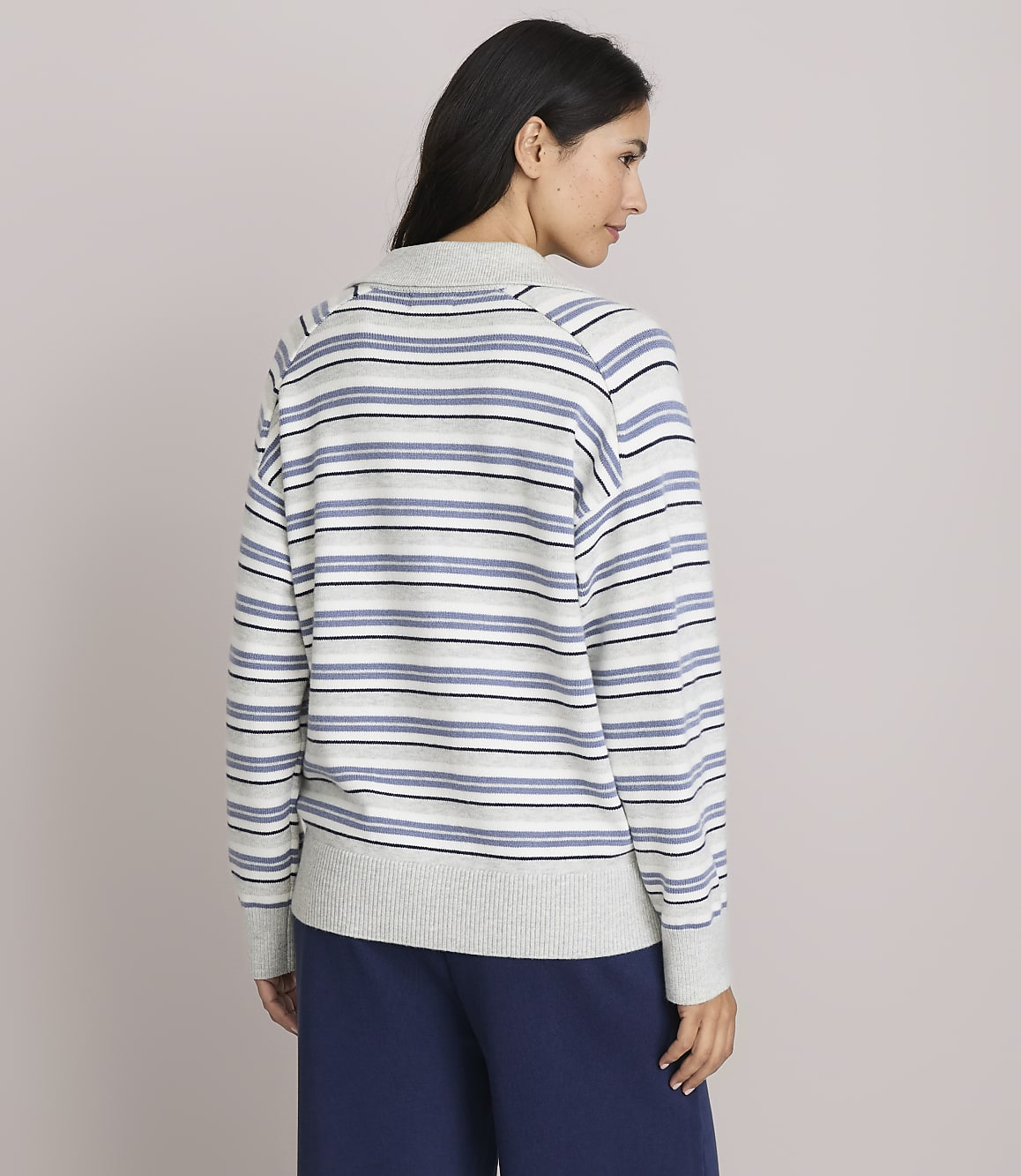A woman in a striped sweater and navy pants stands against a plain, light gray background.