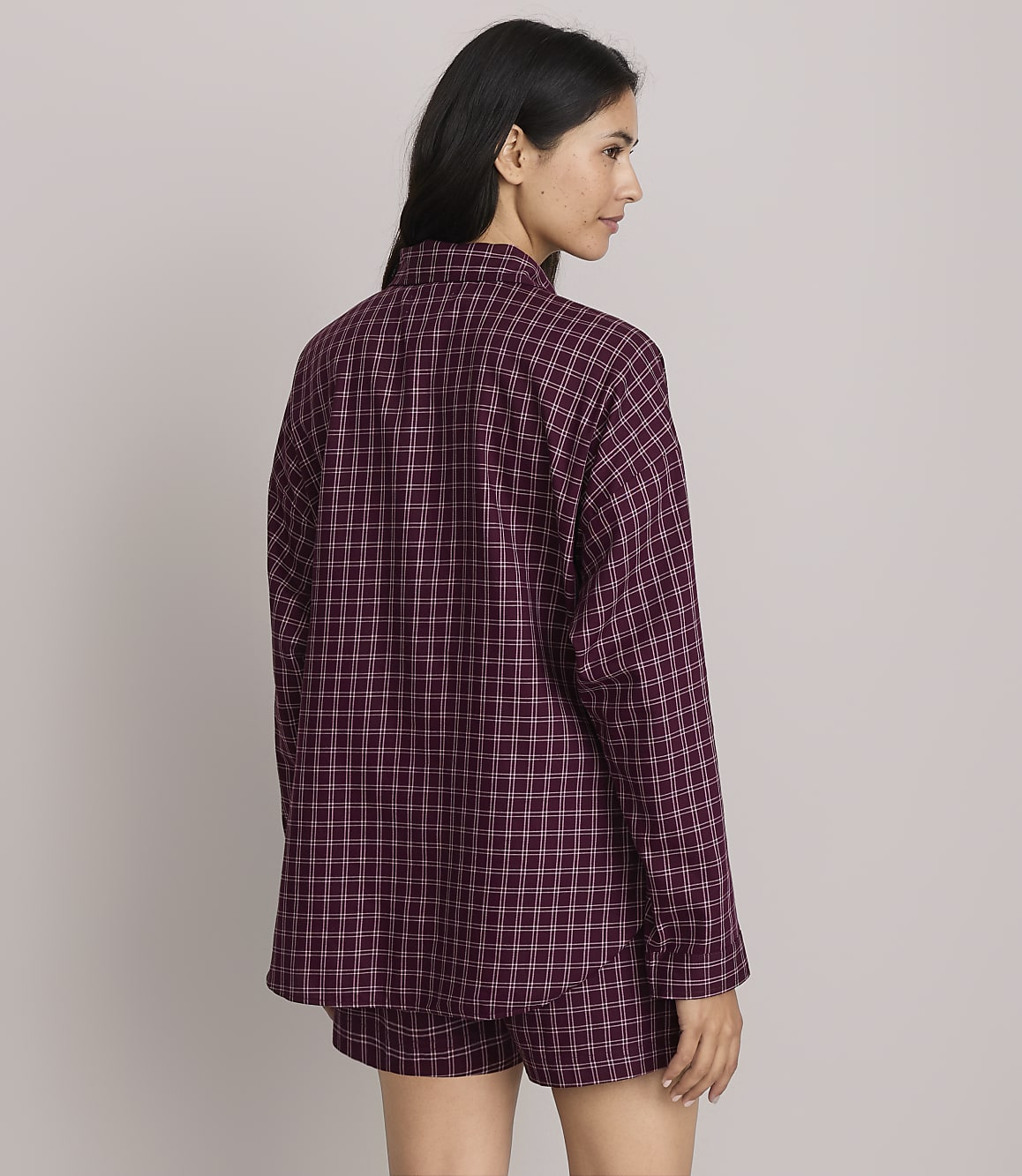 A woman in a burgundy plaid pajama set stands against a plain, light gray background.