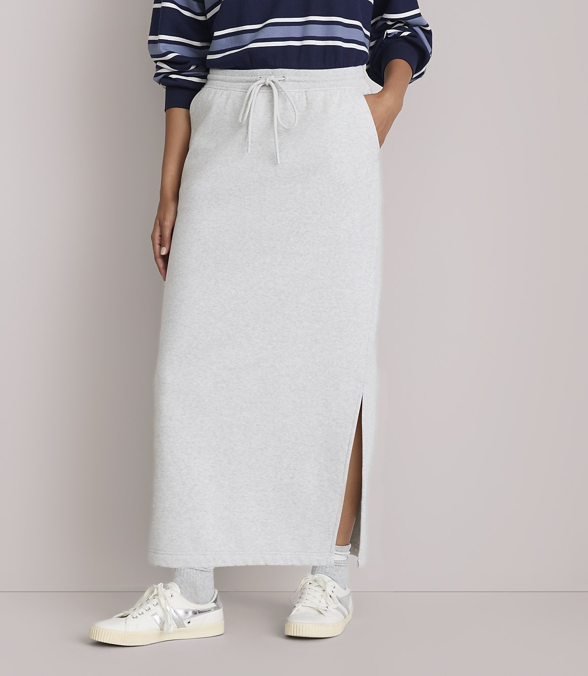 A person stands in a light gray maxi skirt with a side slit, paired with sneakers, against a plain, light-colored background.