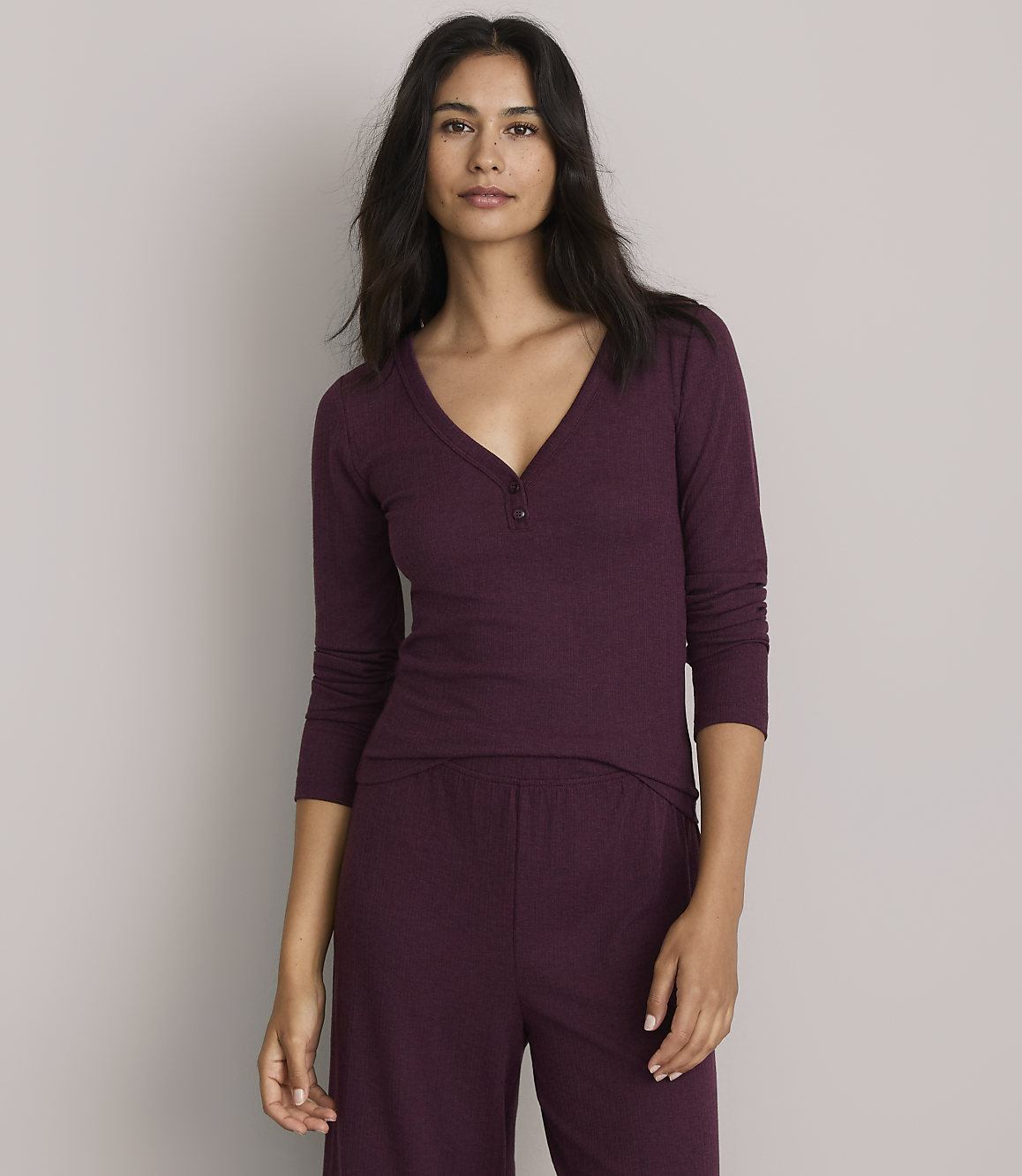 A woman in a burgundy henley shirt and matching pants stands against a plain gray background.